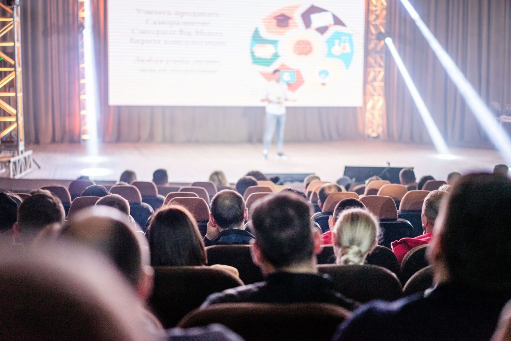 pexels photo 9275222 9275222 1 Audience attentively watching a presentation in a conference setting, with stage lights and speaker visible.